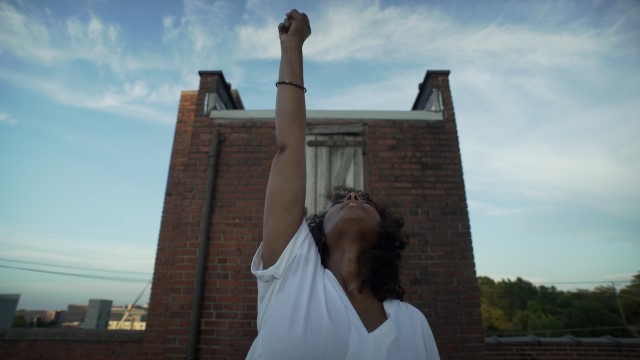 a dark skinned woman in front of a small brick structure raises her face and right arm to the sky