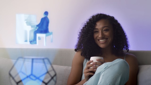 a dark skinned woman smiles warmly while watching a holographic projection of a man playing a piano