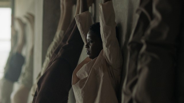 a dark skinned woman raises both arms while pressing her back to a wall