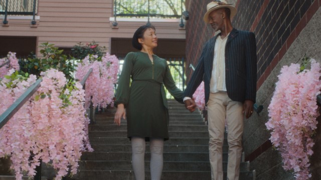 a man and woman hold hands and look lovingly at each other at the bottom of a set of stairs lined with pink flowers