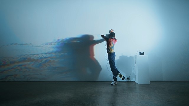 a dancer hooked up to wires faces a wall with a distorted projection of himself on it