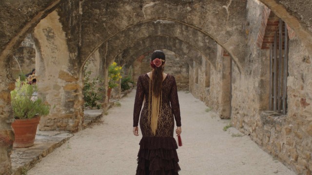 a woman in an elaborate maroon dress walks under a row of stone archways