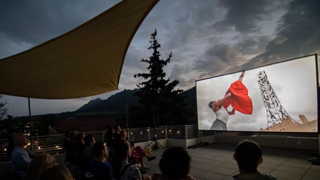 dozens of people watch a dance film on an rooftop at dusk with mountains in the background