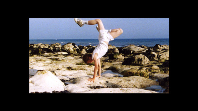 a dancer wearing tennis shorts and shoes does a handstand on large, smooth rocks next to the ocean