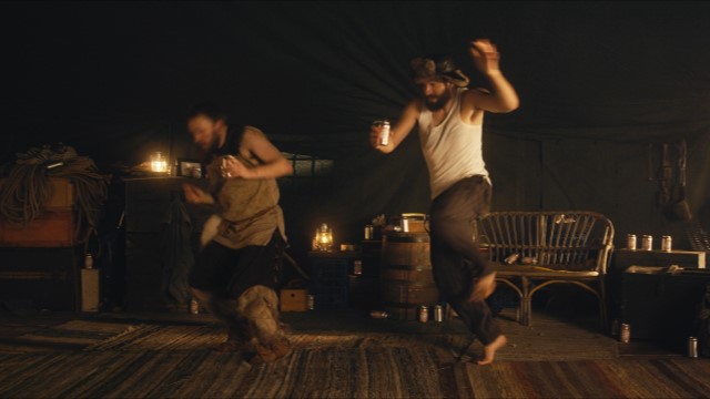 two bearded men in a large, heavy tent dance raucously while holding cans of beer