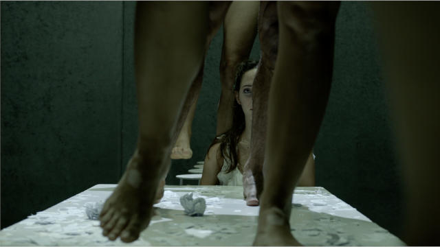a woman looks at the camera, face half obscured by one of five legs standing on tables visible in the foreground
