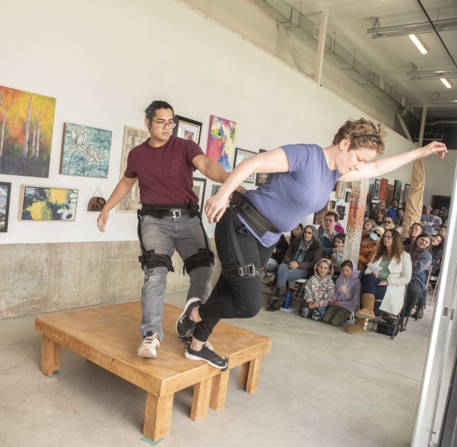 an audience watches a woman stand on the edge of a coffee table, leaning forward while a man behind her holds her by a strap around the waist to keep her from falling