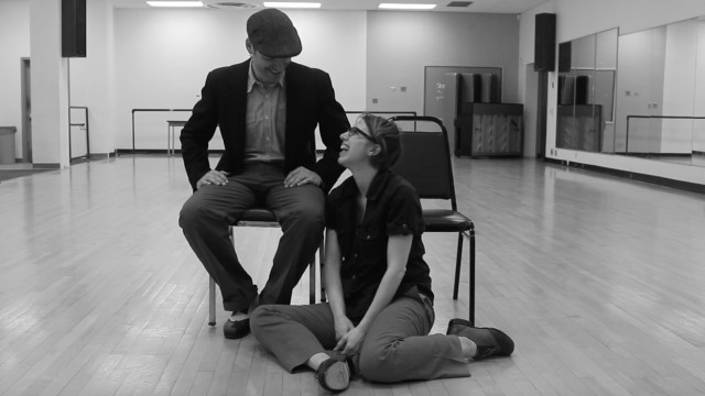a black and white photo of a man in a hat sitting in a chair, laughing with a woman seated on the floor in front of him