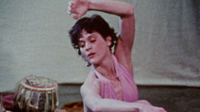 a still frame from old footage of a woman with short hair in a pink dress leaning to her right with a hand drum in the background