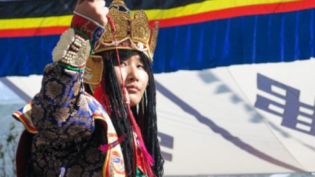 a dancer in an elaborate crown and costume holds up their right hand, looking intently at it
