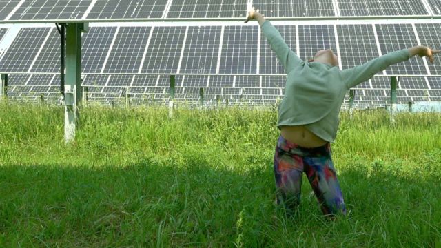 a child throws her arms up in joy while standing in a field of tall green grass under ground-mounted solar panels