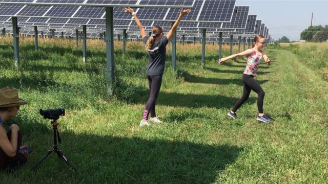 two children dance in a field of solar panels while a dance filmmaker films them