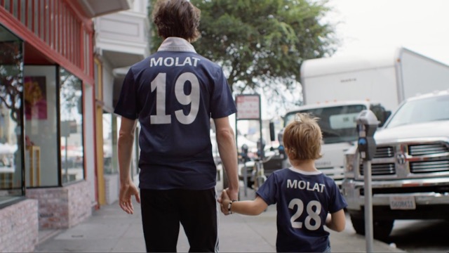 from behind, we see a father and son, holding hands as they walk down a city street, each wearing a soccer jersey with the name MOLAT on the back