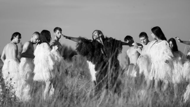 a man in a dark furry robe stretches in a field, surrounded by a group of dancers in white feathered costumes