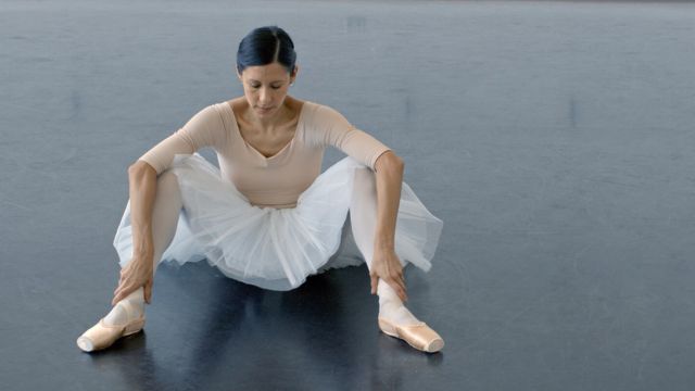 a dark haired woman in ballet clothing sits on the floor, hands resting on her ankles