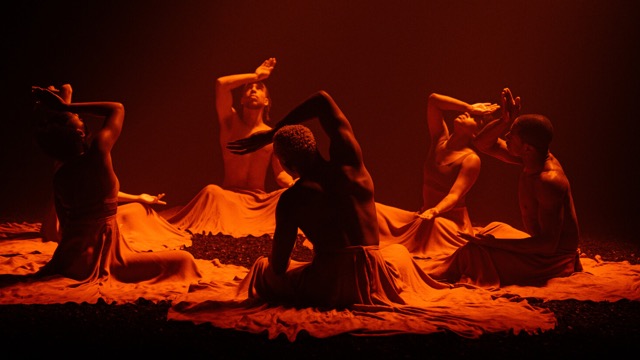 five dancers sit in a circle on the floor of a room under soft, warm lighting