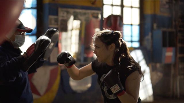 a light skinned woman with long dark hair in a messy ponytail practices boxing with a coach wearing a surgical mask