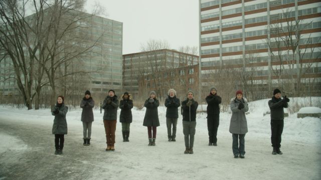 ten dancers in winter jackets stand on an empty ice-covered street with tall buildings behind them