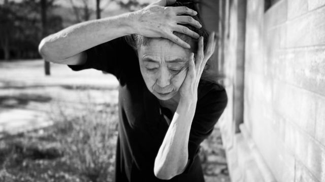 a black and white photo of an older Asian woman with her right hand on her forehead and her left hand covering her left ear