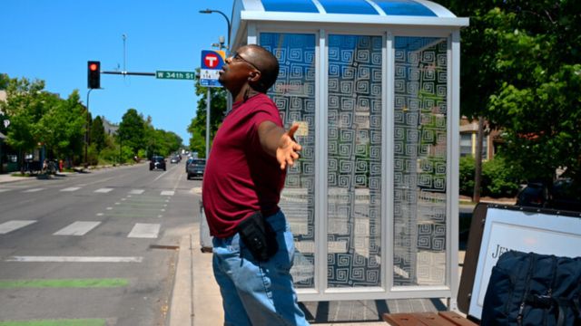 a dark skinned man at a bus stop leans his chest out toward the street