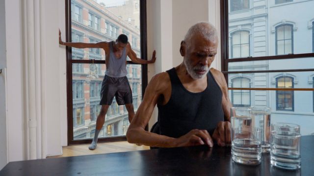 a thin, dark skinned man in a black tanktop sits at a counter next to four glasses of water; behind him, a younger man stretches at a window