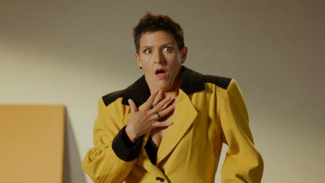 a light skinned dancer with short brown hair and a mustard yellow suit places their hand on their chest and makes a facial expression of shock