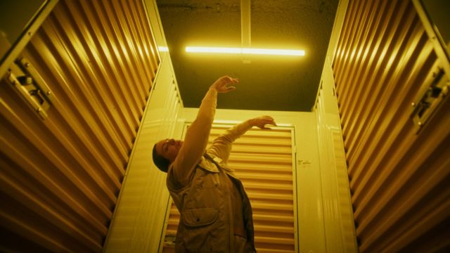 a light skinned woman in a storage facility tilts her head back, arms reaching up