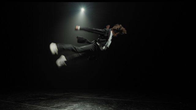 blurred from motion, a dancer in a suit flies across the frame, limbs outstretched