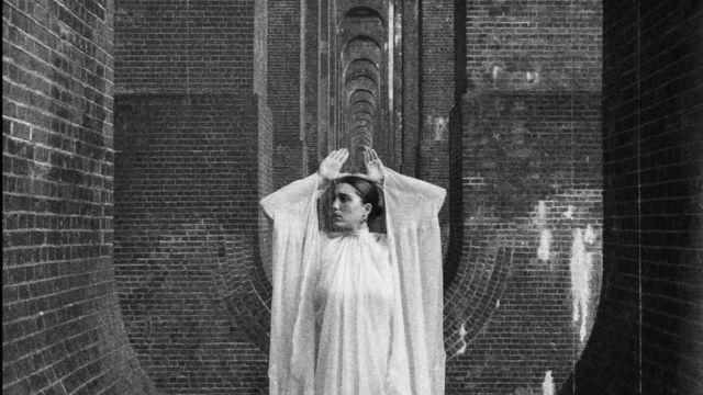 a black and white photo of a woman in a long loose white dress standing in a long hallway made of brick columns