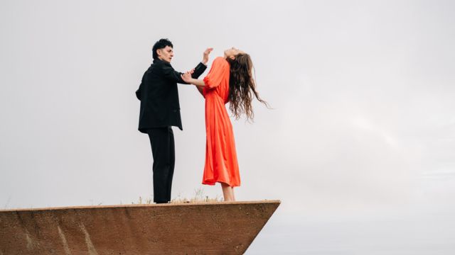 a wide shot of two dancers - one in a black suit and one in a peach dress - stand facing each other near the edge of a roof