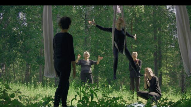 in a forest, a light skinned woman in all black hangs upright in aerial fabric while four other women watch and gesture at her