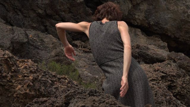 a lean light skinned woman, facing away from the camera, demonstrates lengthy arm movements in a boulder field