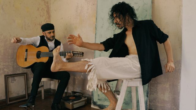 a man with dark, messy hair sits on a stool, left leg extended straight out; behind him a man in a black hat and vest plays an acoustic guitar