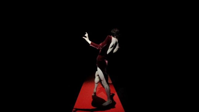 in an otherwise dark room, a dancer in a red velvet dress poses on a strip of brightly lit red carpet