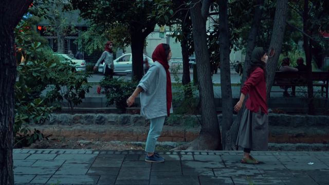 in a public park with trees, two women with face masks lean forward with arms slightly back