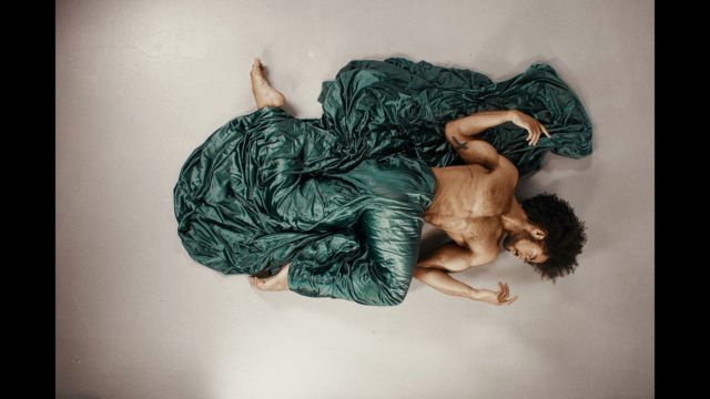 an overhead shot of a shirtless dark skinned man wearing a extremely long dark green vinyl skirt and lying on his back