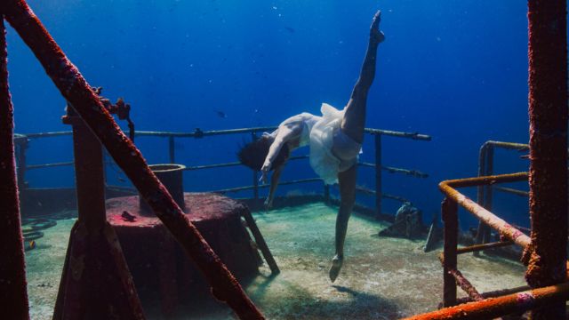 a woman in a white skirt extends her leg upward; she is standing on a shipwreck underwater