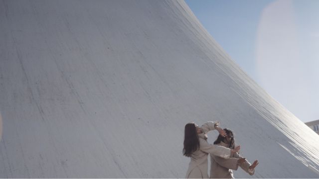 two dancers wearing light colors move in front of a very large, curved white structure that appears to made of adobe or plaster