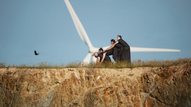 four dancers wearing all black lean on each other on a grassy plateau with a huge wind turbine in the background