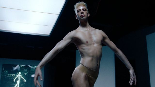 in a room with a famous ballet playing on a screen behind him, a light skinned male ballet dancer with blonde hair faces the camera, naked except for a dance belt