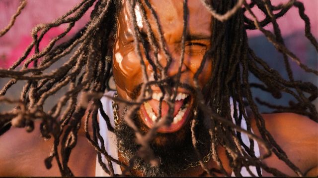 a closeup of the face of a dark skinned man with long, thin dreadlocks, smiling mouth open