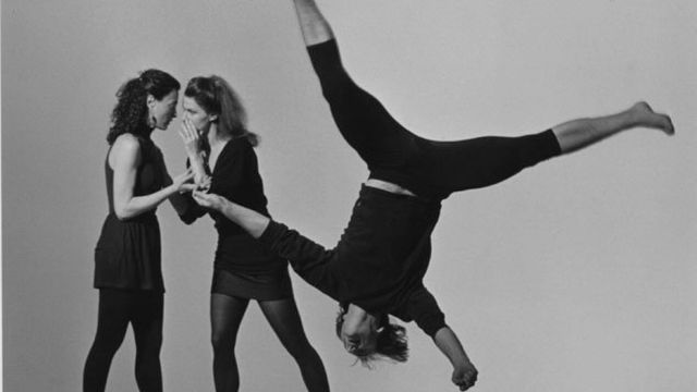 in the background, two women wearing short black dresses talk quietly to each other while another dancer in black cartwheels by in the foreground