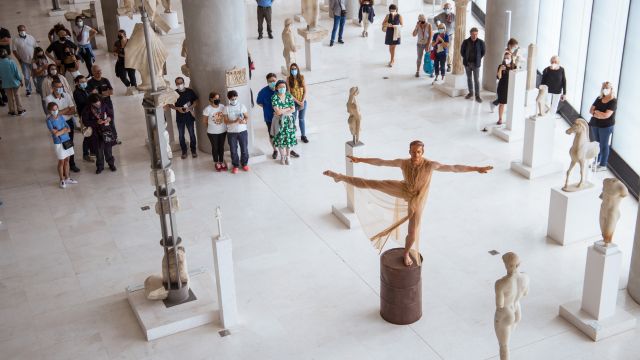 a crowd of people in an art museum watch as a dancer in body paint mimics being one of the sculptures they are surrounded by