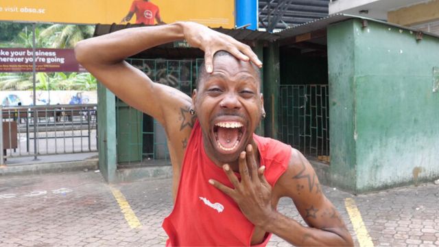 a dark skinned man in a red tank top places his hands above and below his face, mouth open as though screaming