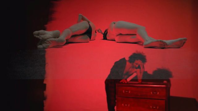 a man leans on a dresser in front of a red wall; two nearly naked people lying on their sides are superimposed on the wall above him