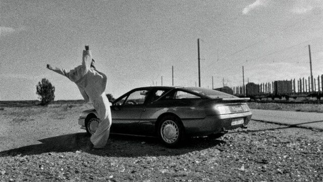 a black and white photo of a dancer wearing baggy white clothes gesturing animatedly at a parked car in a barren landscape