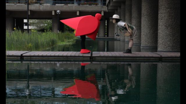 on a walkway over standing water, two people facing each other bend at the waist, their heads close together; one is wearing a blocky red bird costume