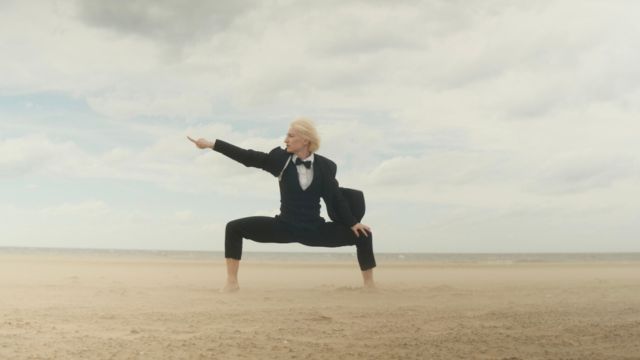 a light skinned woman with short blond hair wearing a black tuxedo squats powerfully on a vast landscape of sand and points off frame