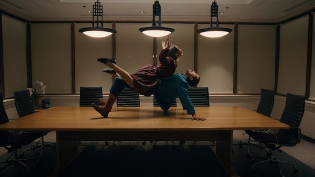 a woman balances on a man's pelvis on top of a large table in an empty conference room