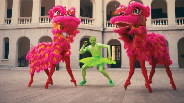 a ballet dancer in green body paint and green tutu poses between two sets of ballet dancers en point wearing extravagant pink dragon costumes